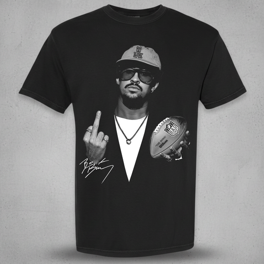 A black short-sleeve graphic t-shirt featuring a black and white print of a man wearing a cap and sunglasses, holding a football and making a middle finger gesture, with a signature graphic below. The t-shirt is displayed against a textured gray background.
