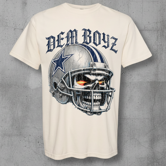 An off-white graphic t-shirt featuring a Dallas Cowboys skull helmet design with the text 'DEM BOYZ' above it, displayed against a textured gray concrete background.