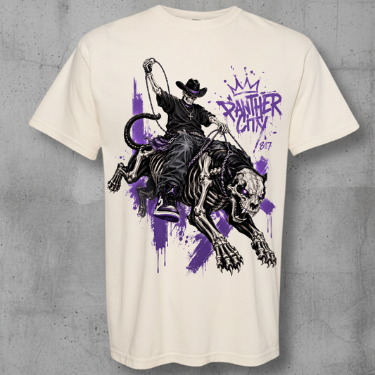 An off-white short-sleeve t-shirt with a graphic print of a skeleton cowboy riding a black tiger, set against a purple paint splatter background with the text 'panther city ' and a crown. The t-shirt is displayed flat against a textured gray concrete wall.