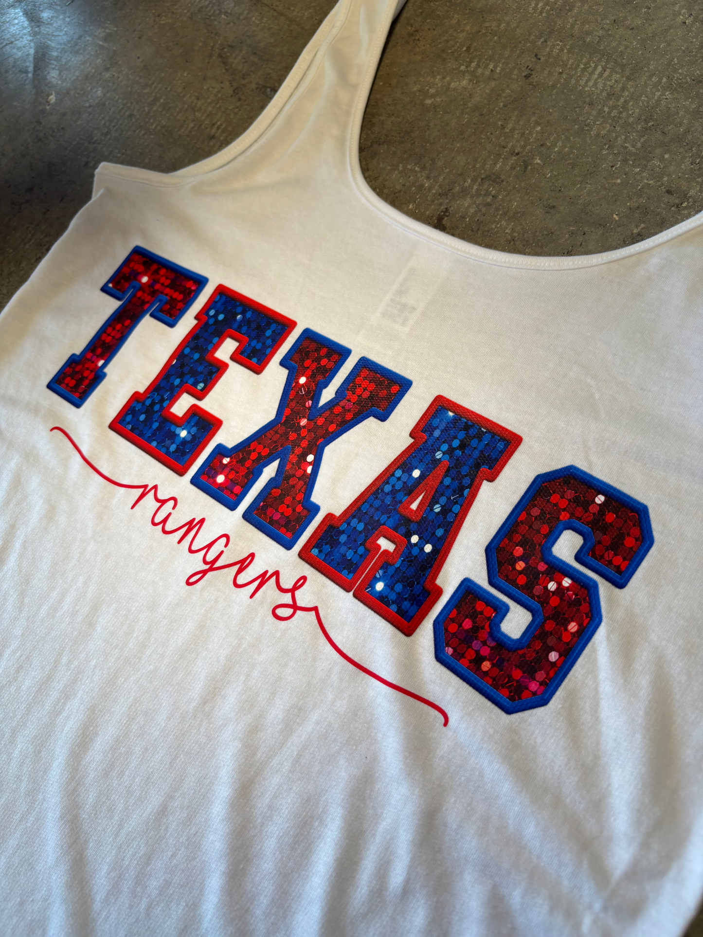 White tank top with 'TEXAS' in red and blue sequins on a gray surface