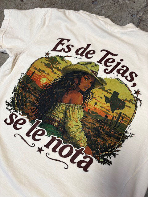 White t-shirt with a graphic design of a woman in a hat and text 'Es de Tejas, se le nota' on a textured surface.